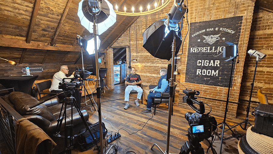 Two men are seated in a recording studio with professional lighting and cameras, preparing for a podcast interview at